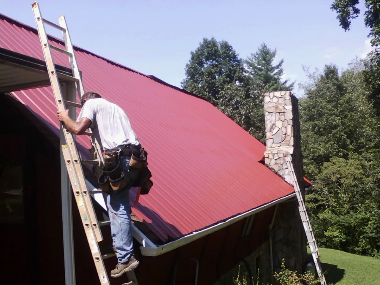 Roofer on a ladder beside a red metal roof and stone chimney — Soffit & Fascia Repair by Roofing Brothers in Nephi